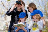 Eltern mit Kinder bei der Vogelzählung. Vater mit Fernglas - 2 Kinder und die Mutter schauen interessiert in einem Nachschlagswerk (Buch)